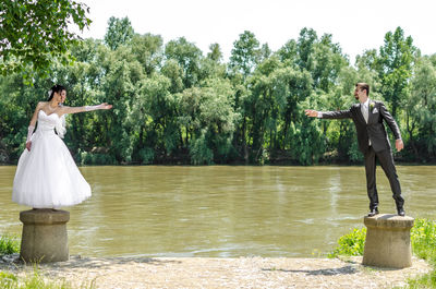 Full length of wedding couple gesturing while standing on bollard against river