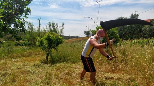 Side view of man removing grass on field against sky