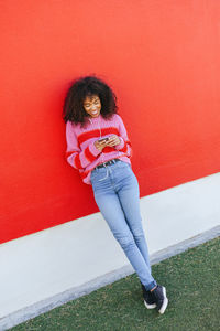 Smiling young woman with earphones leaning against red wall looking at cell phone