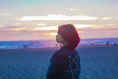 Rear view of woman with dyed hair while standing at beach against sky during sunset