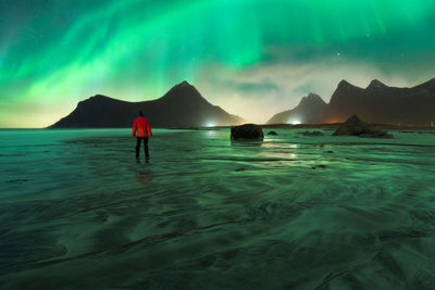 Back view of unrecognizable male tourist admiring snowy mounts under bright aurora borealis lights on lofoten island