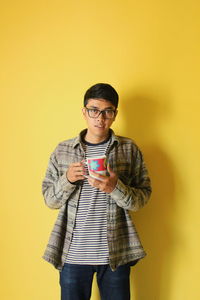 Portrait of man wearing eyeglasses holding coffee cup standing against yellow background