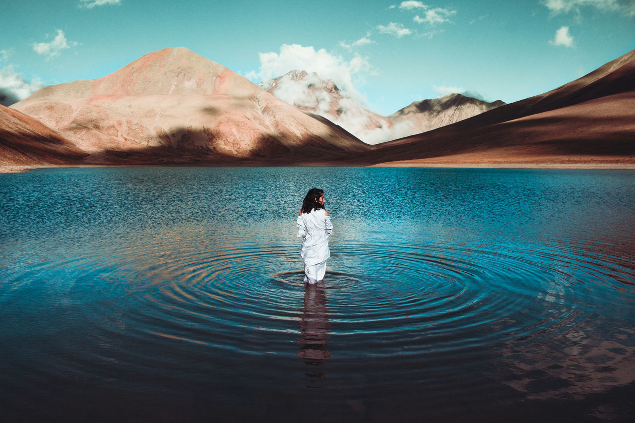 REAR VIEW OF MAN STANDING IN LAKE AGAINST SKY