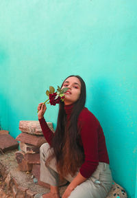 Portrait of young woman holding rose while crouching against wall