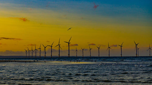 Wind turbines by sea against sky during sunset