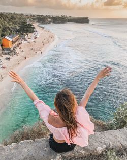 High angle view of carefree woman with arms raised sitting on retaining wall over sea