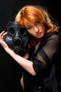 Portrait of young woman holding gas mask against black background