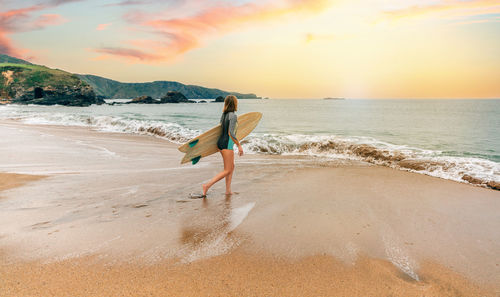 Surfer woman with wetsuit carrying surfboard looking to the sea at the beach