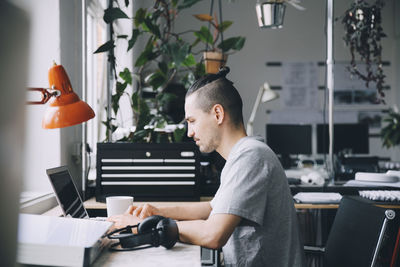 Side view of male hipster using laptop at desk in creative office