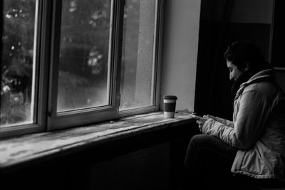 Side view of smiling woman using phone by window at home