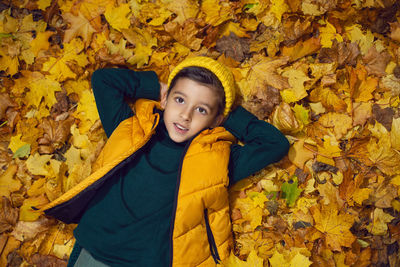 A boy child in a yellow jacket and hat lies on autumn leaves in the forest