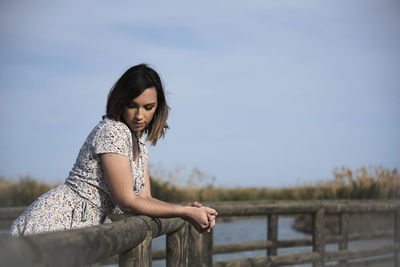 Young woman looking away while sitting on railing against sky