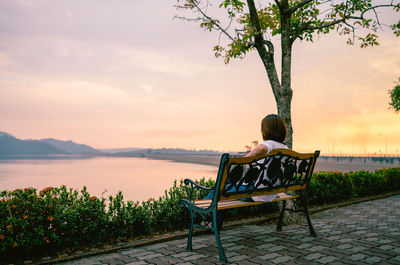 Rear view of mature woman looking at lake while sitting on bench against sky during sunset