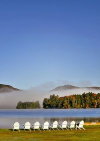 Empty chairs on grassy field by lake against clear sky during foggy weather