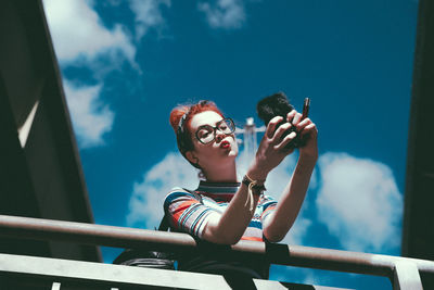 Low angle view of young woman taking selfie with mobile phone while leaning on railing against sky