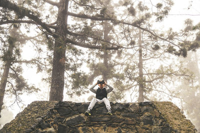 Full length of boy looking through binoculars while sitting on retaining wall at forest