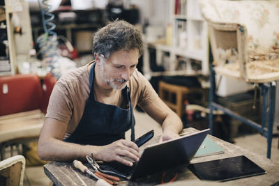 Male upholstery worker using laptop while sitting at workbench in workshop