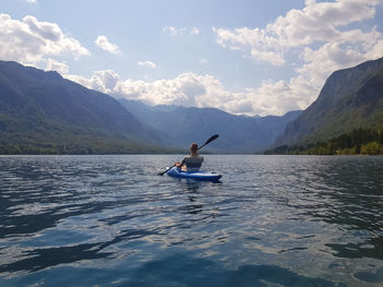 Rear view of man canoeing on lake by mountains against cloudy sky