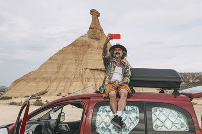 Traveler sitting on top of a van using a mobile camera to take one with the landscape 