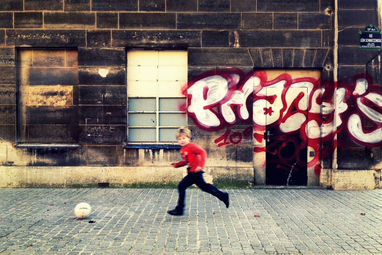 full length, building exterior, built structure, street, architecture, graffiti, sidewalk, walking, childhood, lifestyles, casual clothing, text, wall - building feature, outdoors, side view, standing, day, city
