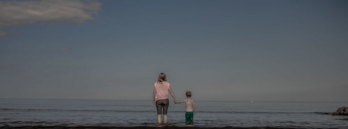 Rear view of mother and son standing in sea against sky at coldingham bay