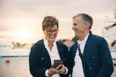 Happy senior woman using smart phone while standing by man at harbor