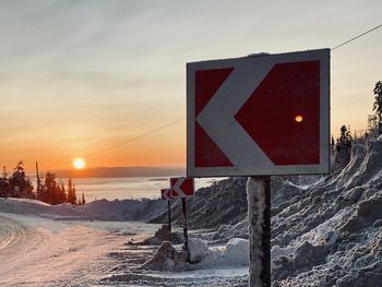 Road sign on snow covered field against sky during sunset