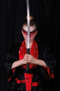 Portrait of young woman wearing kimono and make-up holding sword against black background