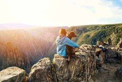 Girl sitting with dog on rocks against mountains during sunny day