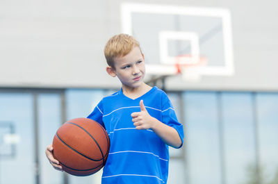 Thoughtful boy gesturing thumbs up while holding ball against basketball hoop
