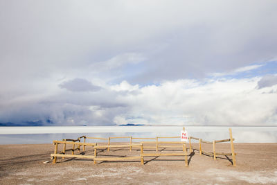 Wooden fence by salt flats against cloudy sky on sunny day