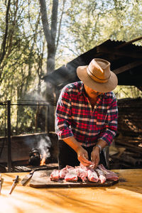 Adult argentinian male with mustache wearing beige hunter hat and checkered shirt salting meat on cutting board while preparing grill in picnic in nature