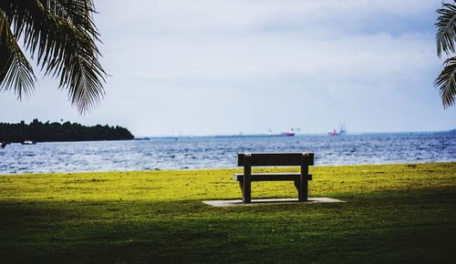 Empty bench on grass by sea against sky