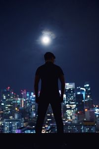 Rear view of silhouette man standing on building terrace against illuminated cityscape at night