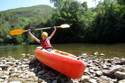 Excited woman holding oar while sitting on kayak at riverbank