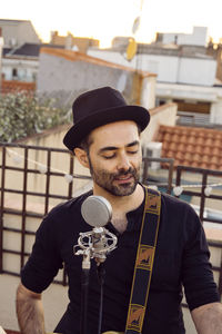 Portrait of young man wearing hat singing by microphone standing against sky