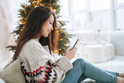 Young asian woman in cozy sweater using mobile smartphone on bed in room with christmas tree