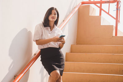 Full length of young woman using phone while standing against wall