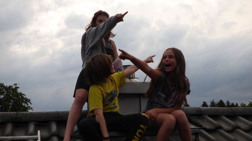 Low angle view of siblings pointing while sitting on roof against cloudy sky