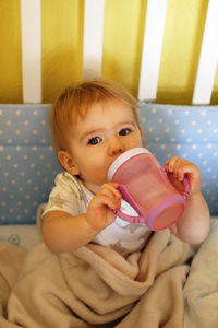 Cute one year old girl is drinking water from bottle after waking up in the morning in her bed.
