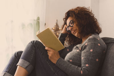 Woman reading book while relaxing on sofa in living room at home