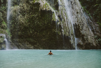 Rear view of shirtless man swimming in river by waterfall at forest