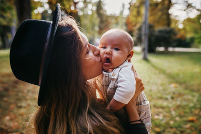 Loving young mother in hat hugging and kissing cute little baby while resting on grassy meadow in park