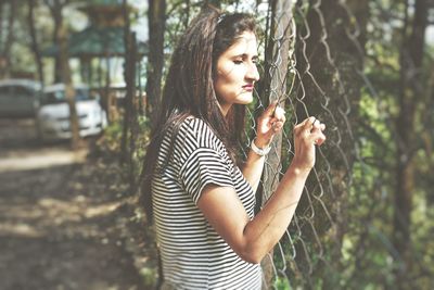 Young woman looking through fence at park during sunny day