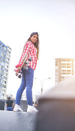 Beautiful young woman in cap is sitting at skatepark on the ramp with her longboard.