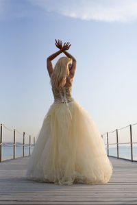 Rear view of young woman wearing evening gown standing on pier against sky