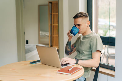 A young man drinks coffee and works remotely on a laptop while sitting at a table in the living room