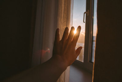 Cropped hand reaching towards window at home during sunset
