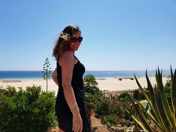 Side view portrait of smiling woman wearing sunglasses standing at beach against sky during sunny day