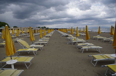 Closed parasols and lounge chairs at sandy beach against cloudy sky